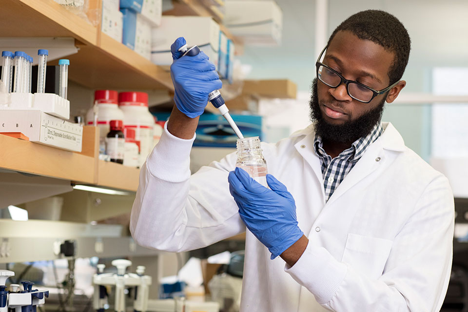 Graduate student in a laboratory setting shown pipetting liquid into beaker.