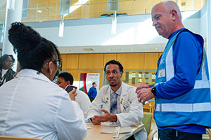 Two student pharmacists seated at a table in white coats, participating in a POD drill. A volunteer in a blue vest is standing at the end of the table.