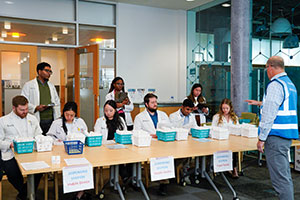 Group of PharmD students in white coats, seated at a long table with baskets of medications in front of them. The students are participating in a POD drill.