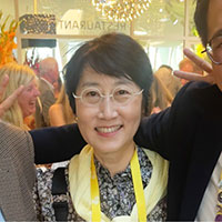 Sandy Rhie posed indoors at a celebratory event, wearing a yellow scarf and glasses.