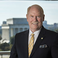 Thomas Menighan posed outdoors, wearing a dark suit jacket and a yellow tie, with buildings in the background.