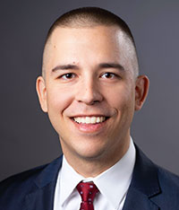 Portrait of Ryan Constantino, wearing a navy jacket, posing against a dark gray background.