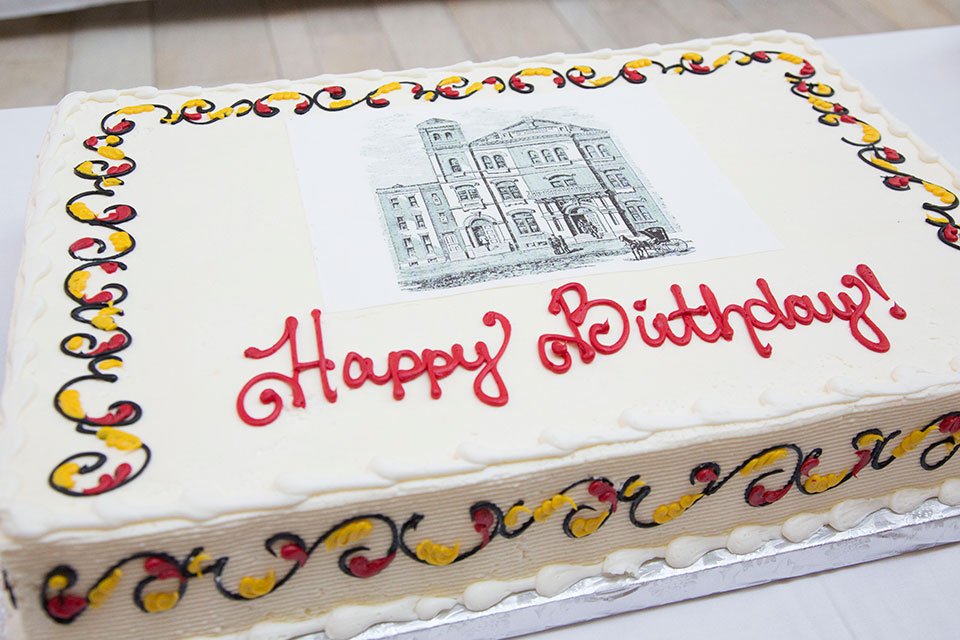 White sheet cake with red and yellow flourishes that says happy birthday with a red and white building pictured.