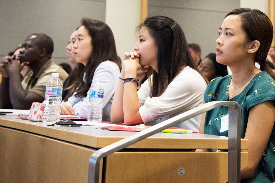 Students sit at tables in lecture hall attentively listening to the dean speak.