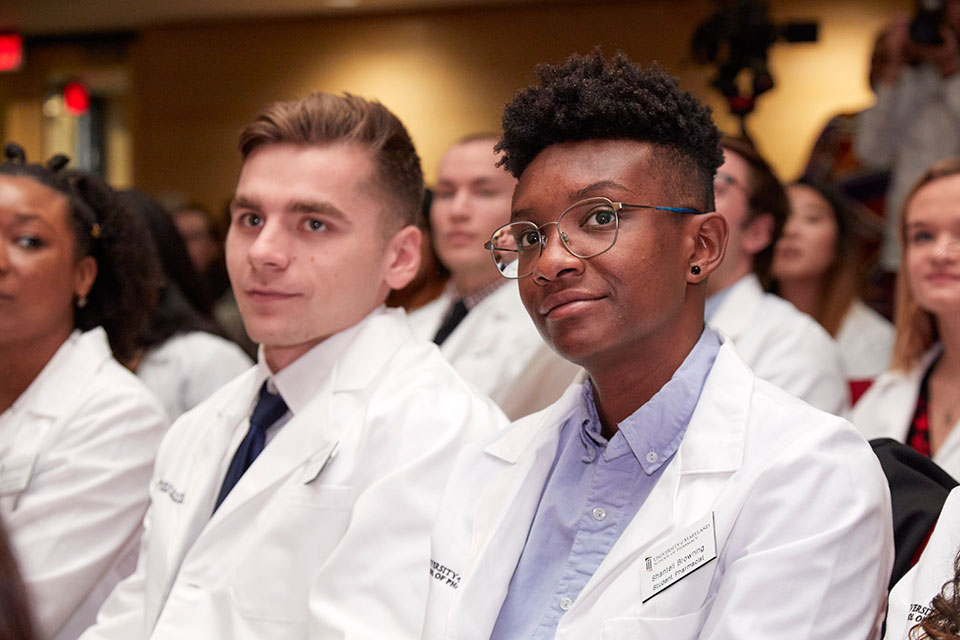 Student pharmacists donning their white coats sit in the audience and gaze at the stage.
