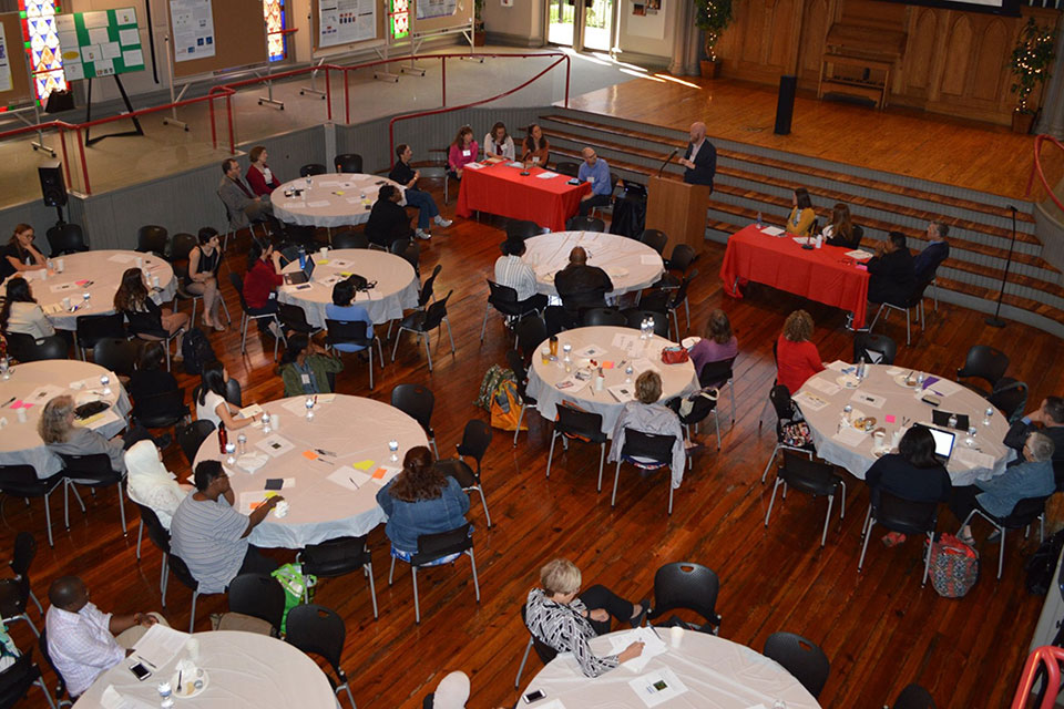 Overhead photo of conference attendees seated at tables listening to a presenter.