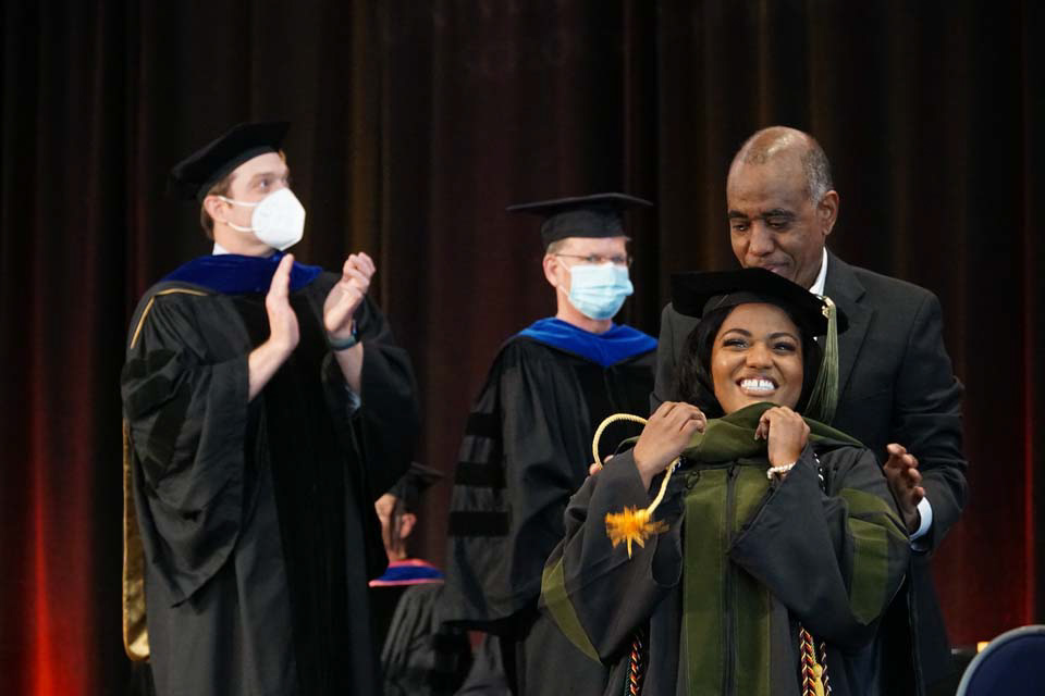 A student smiles as she is hooded by her father on stage