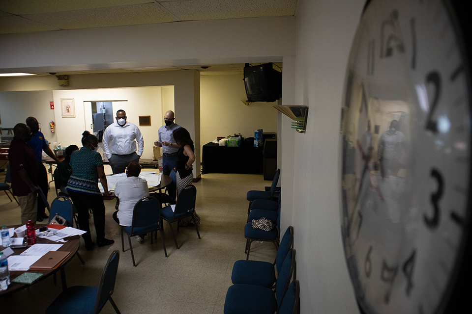 A group of people huddle around a table. A clock is prominently featured in the foreground on the right side of the image.