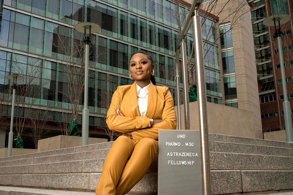 Anyen Fon sits on steps in front of an office building with a sign that says UMD PharmD, MSc, AstraZeneca fellowship.
