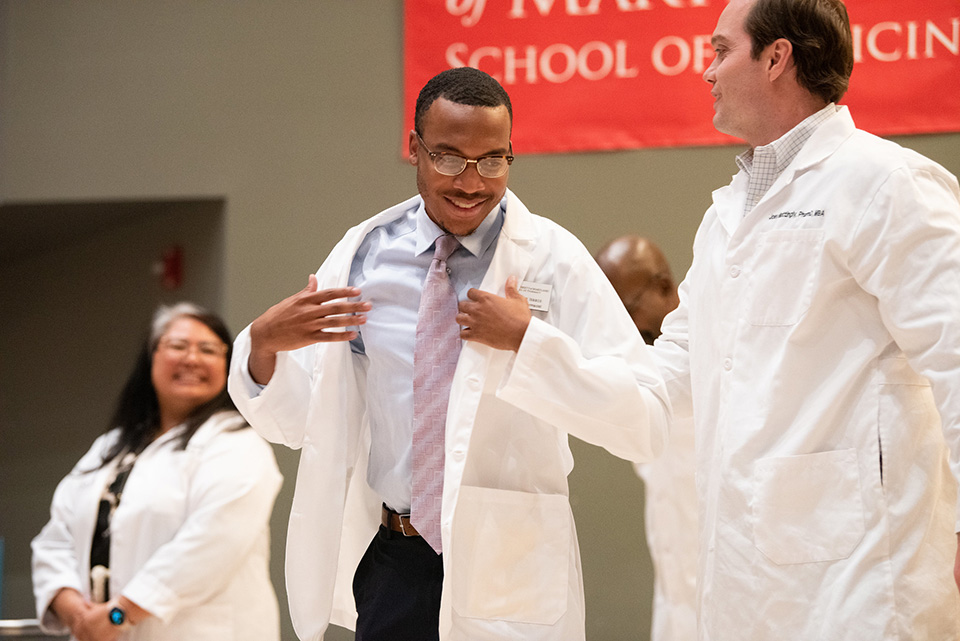 A student adjusts his white coat next to faculty.
