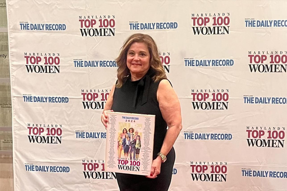 Magaly Rodriguez de Bittner holding a Maryland Top 100 Women sign and standing in front of a Maryland Top 100 Women backdrop.