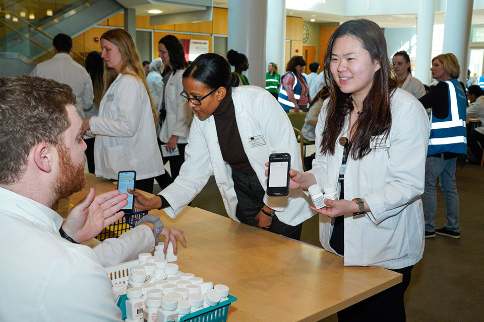 A group of third year pharmacy students in white coats participating in a POD drill in Pharmacy Hall. One group of students is standing on one side of a long table, and another group is seated at the table.