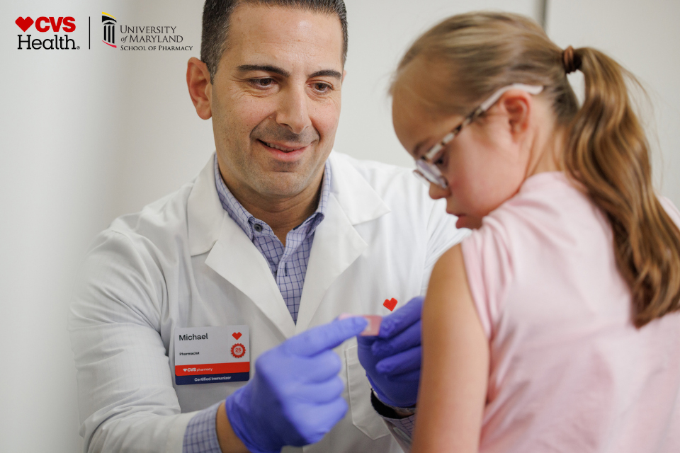 Pharmacist placing a bandage on a patient’s arm with CVS Health and UMD School of Pharmacy logos