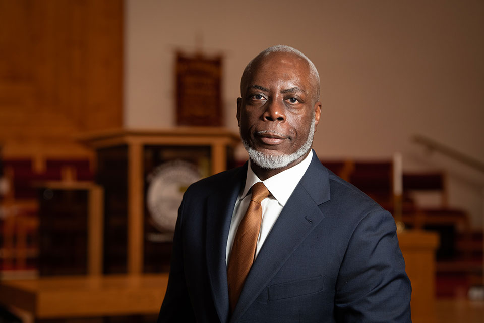 Dr. Franklin Lance in a dark suit and tie standing indoors in front of a church pulpit. The background includes wooden furnishings and softly lit seating.