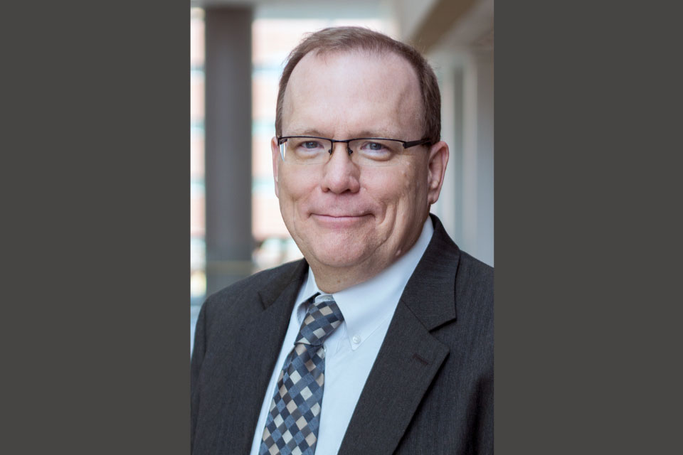 Portrait of Bill Cooper wearing a dark suit jacket, a white dress shirt, and a patterned tie, standing indoors with a blurred hallway background