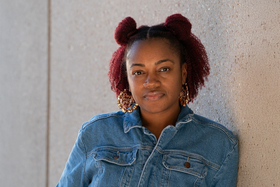 Employee of the Month Paris Barnes poses for a portrait wearing a denim jacket and large earrings, standing against a concrete wall.