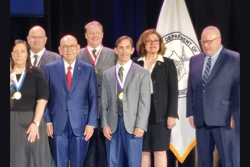 Daniel Mullins, centered, poses with fellow recipients at an award presentation, with medals visible and a department banner in the background