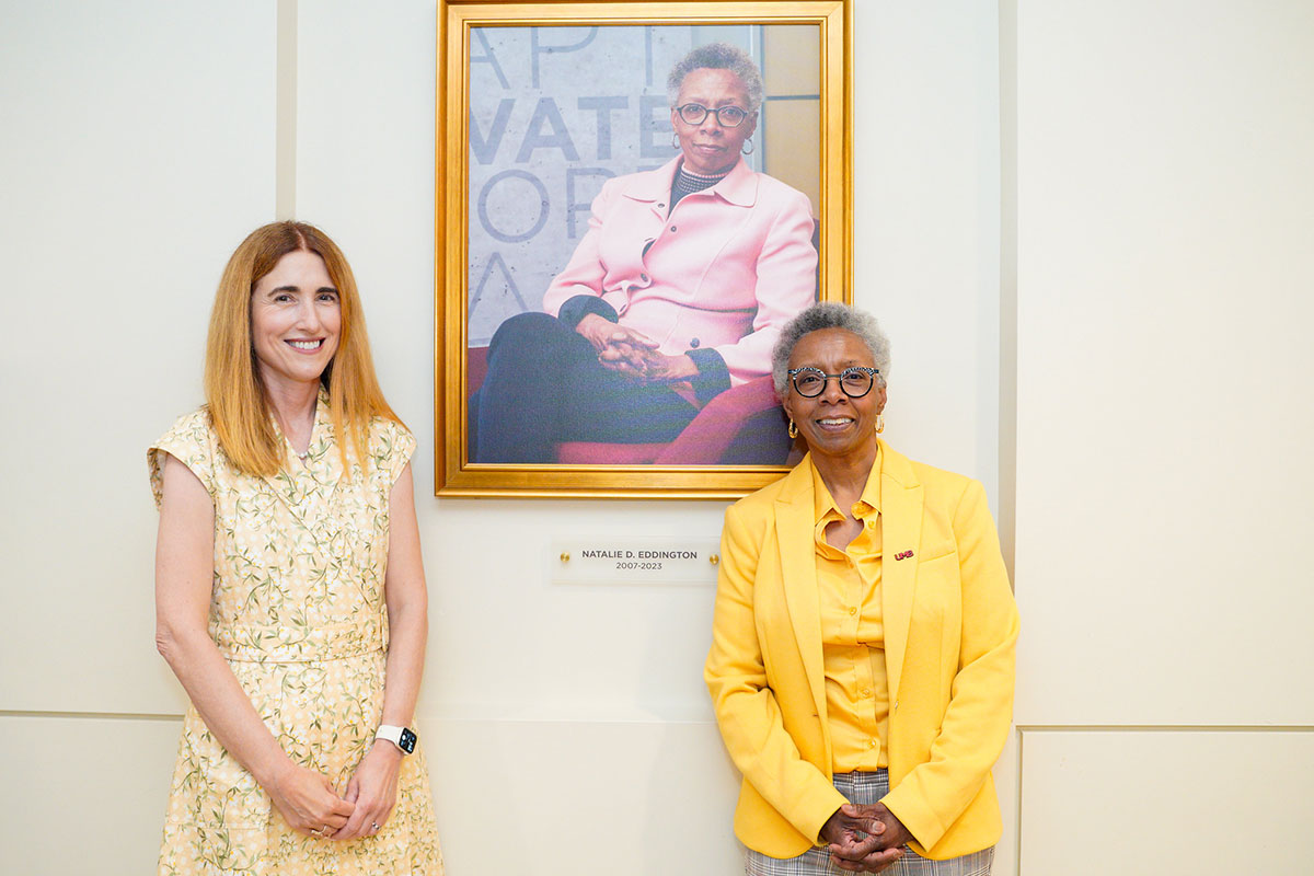 Dean Sarah Michel and Dean Emerita Natalie Eddington pose beside a framed portrait honoring Natalie Eddington in Pharmacy Hall.