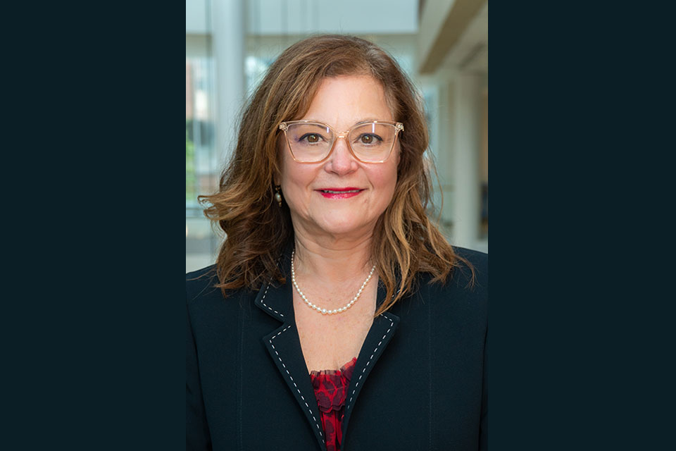 Magaly Rodriguez de Bittner, wearing a dark blazer and light glasses, posed inside with a blurred background of windows.