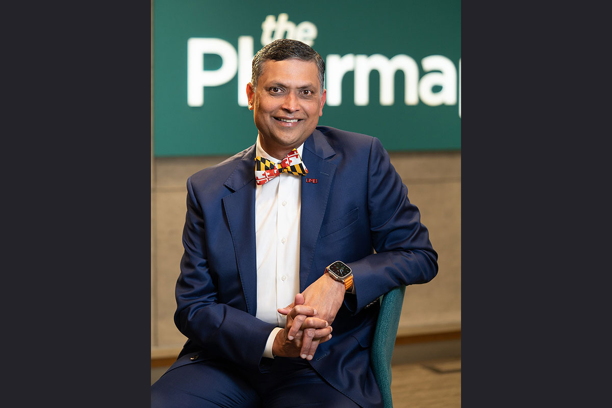 Portrait of Joga Gobburu seated indoors, wearing a blue suit and Maryland‑themed bow tie, with a School of Pharmacy backdrop.