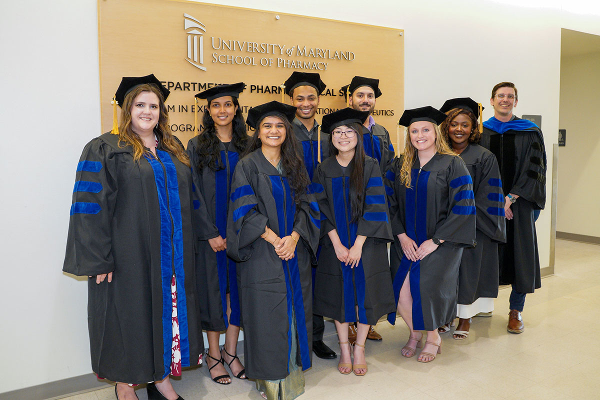 PhD in PSC graduates in caps and gowns stand with Ryan Pearson in front of a University of Maryland School of Pharmacy sign indoors.