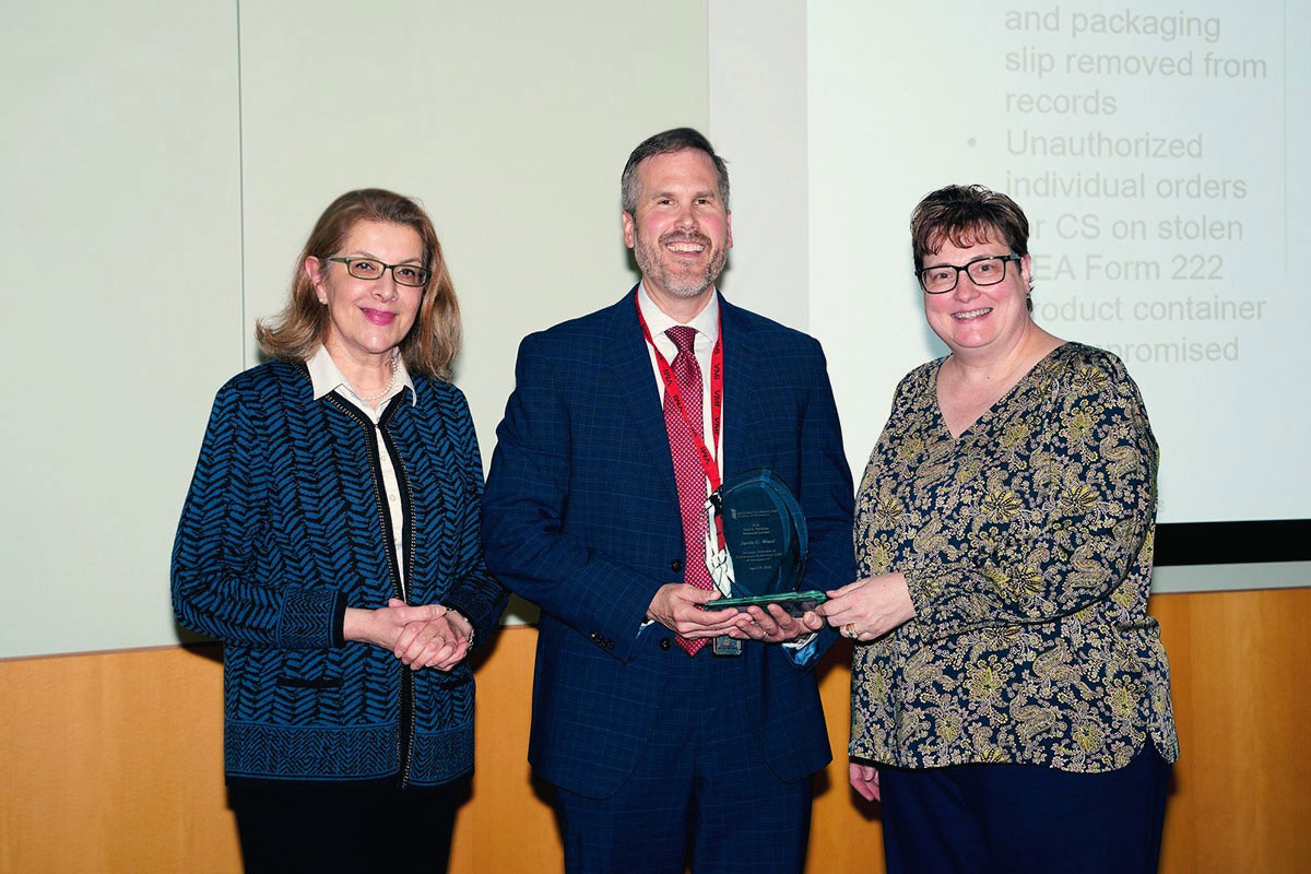 Fadia Shaya, Justin Wood of the DEA, and Jill Morgan stand together as Wood holds an award following his presentation.