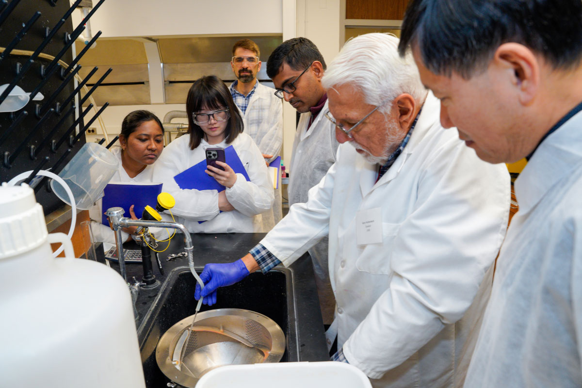 Participants in the Tablets and Capsules short course, all wearing white coats, observe an instructor demonstrating a lab technique over a sink in a classroom laboratory.